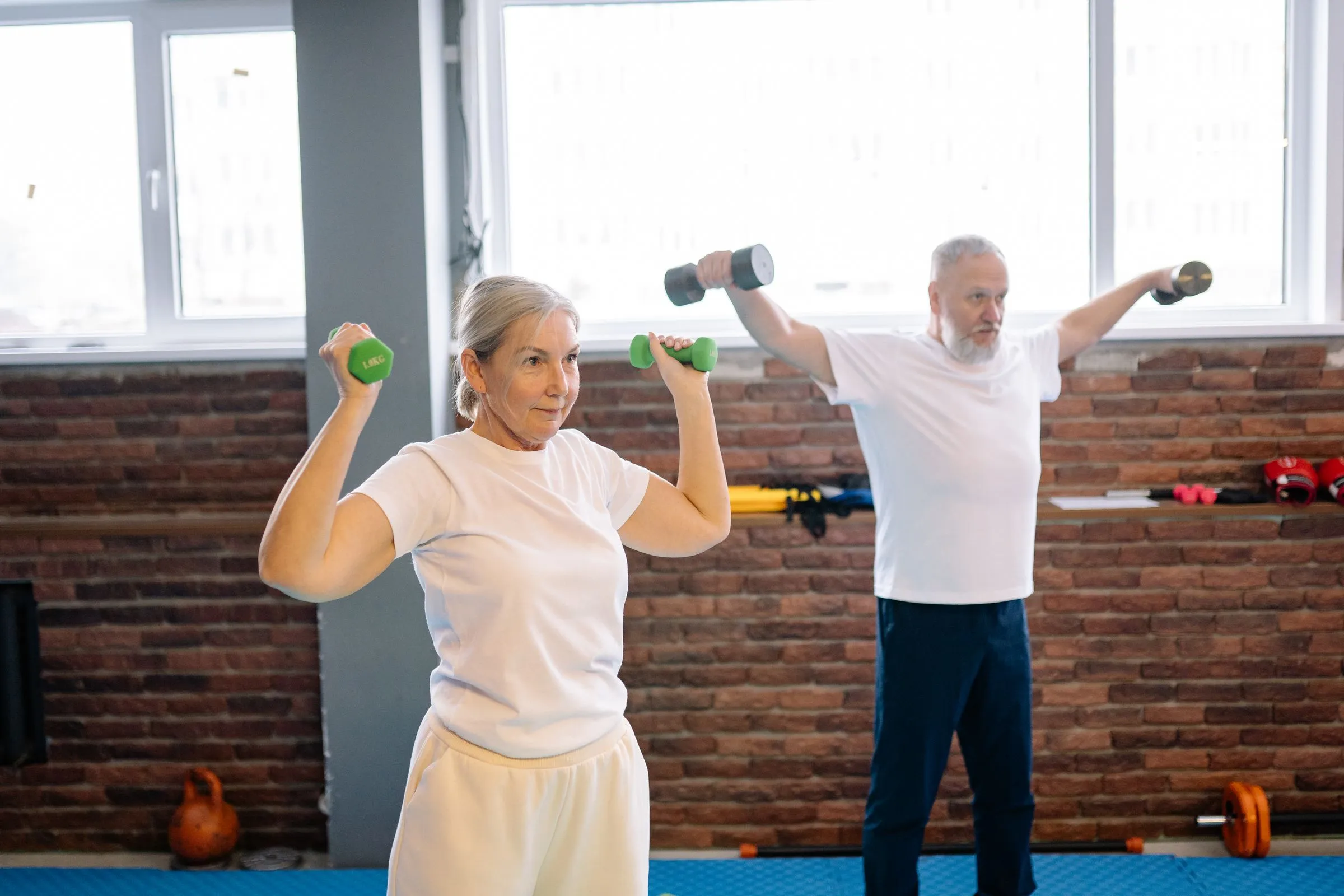 An older couple lifting light dumbbells overhead together in a bright gym.