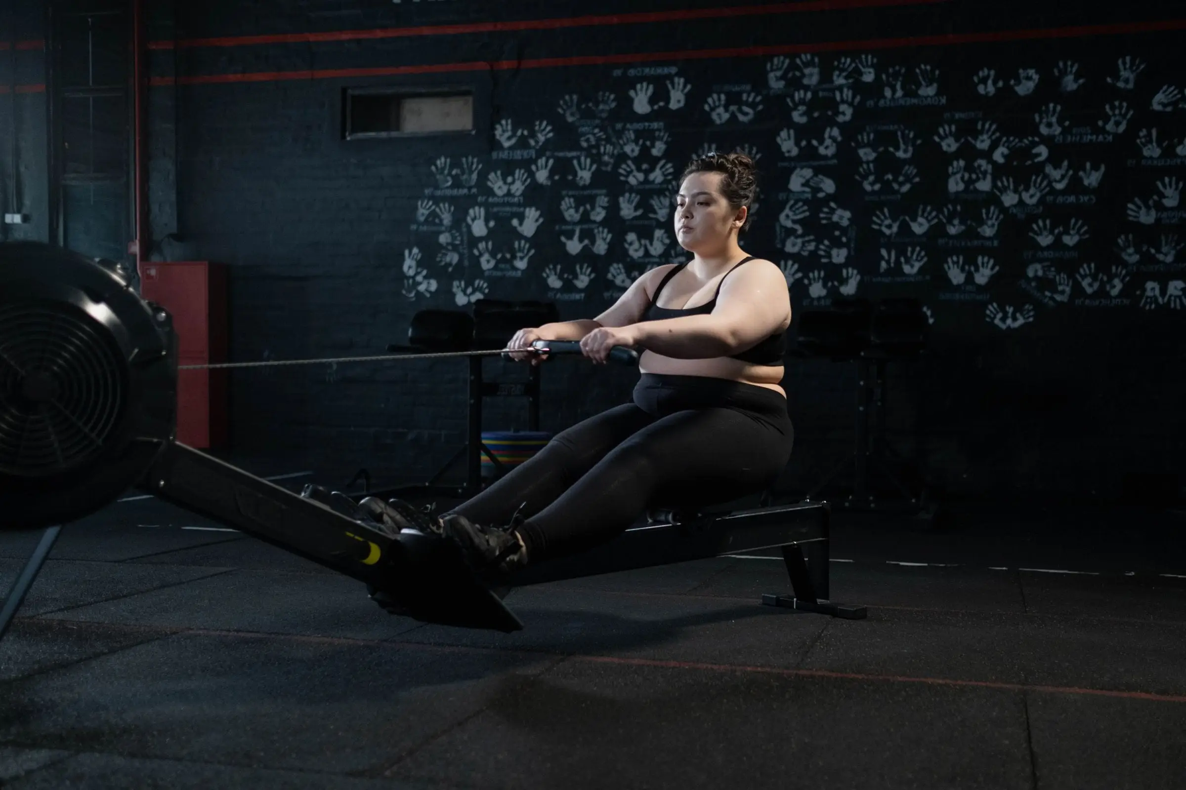 A member mid-stroke on a rowing machine in a dark gym with a rubber floor and a wall of chalk handprints.